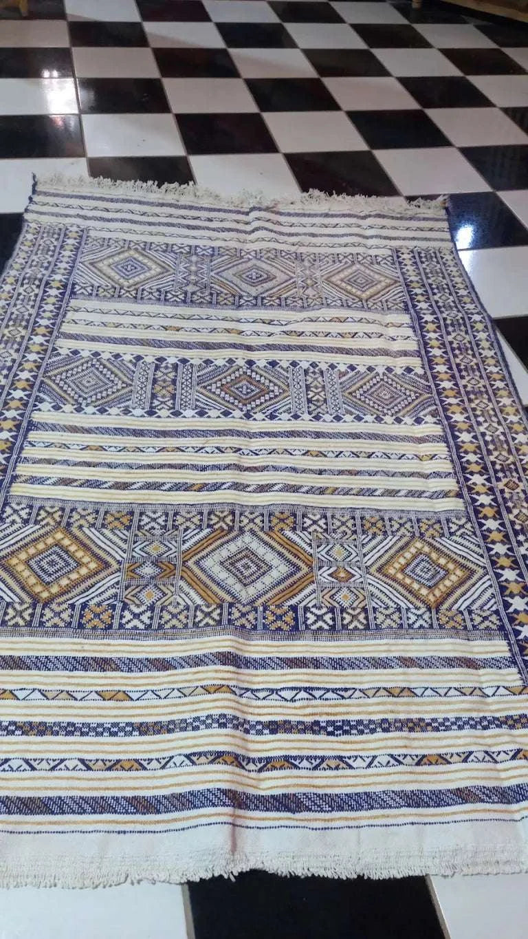 Moroccan woven rug with geometric patterns in blue, brown, and white colors displayed on a checkered floor at Dépôt Nomade.
