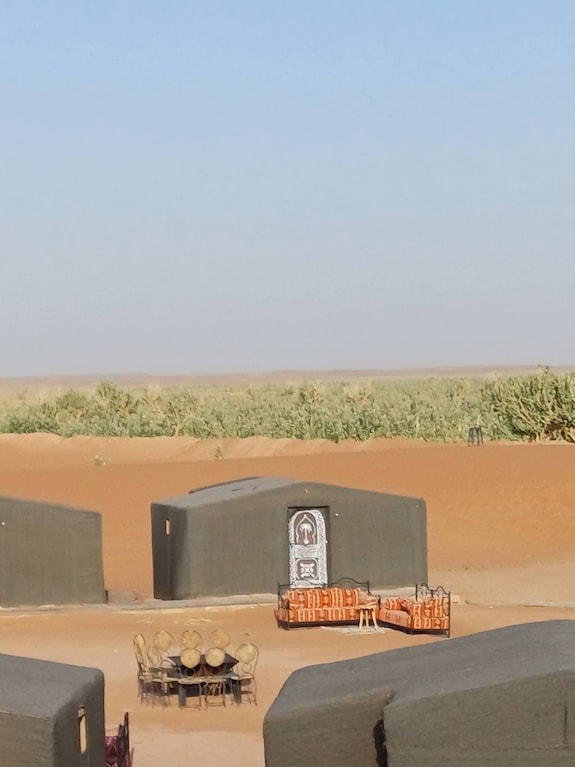 Desert Erg Chigaga Camp with traditional black Berber tents arranged around outdoor seating and tables against sandy dunes and clear sky