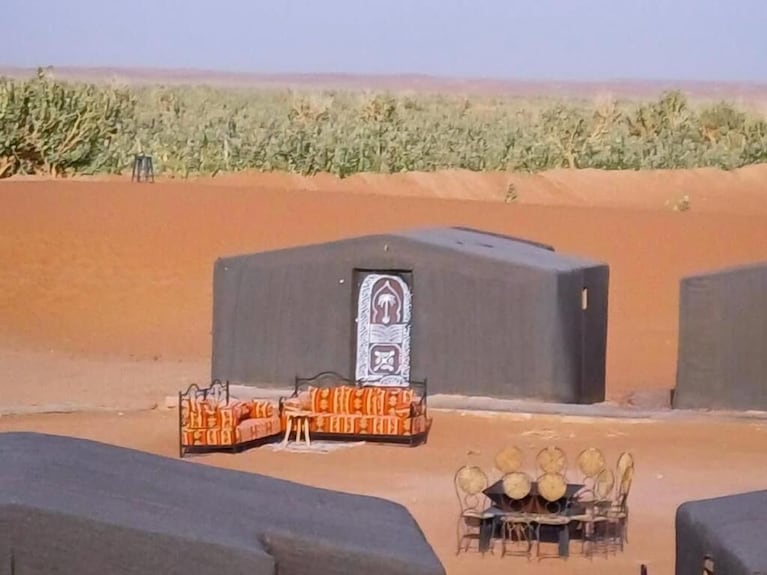 Desert Erg Chigaga Camp with black fabric tents and orange patterned seating area on sand with greenery in background