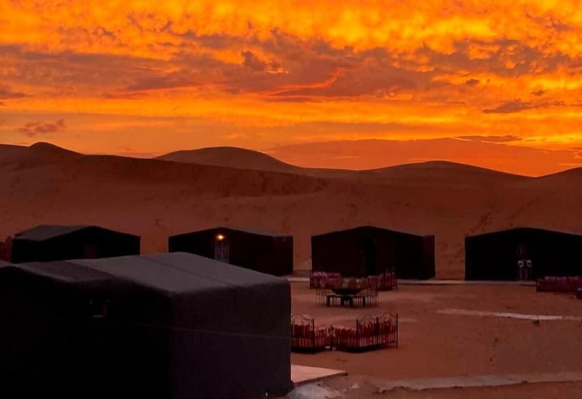 Desert Erg Chigaga Camp with black Berber-style tents set against orange and yellow sunset skies over sand dunes