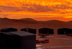 Desert Erg Chigaga Camp with black Berber-style tents set against orange and yellow sunset skies over sand dunes