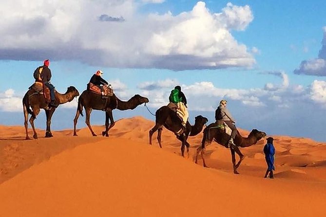 Desert Erg Chigaga Camp camel trek with riders in colorful clothing crossing golden sand dunes under blue sky with clouds
