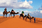 Desert Erg Chigaga Camp camel trek with riders in colorful clothing crossing golden sand dunes under blue sky with clouds