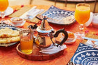 Traditional Moroccan tea set with silver teapot, glasses of orange juice, blue patterned plates, and pastries on an orange tablecloth at Desert Erg Chigaga Camp