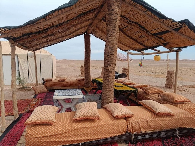 Desert Erg Chigaga Camp outdoor seating area with patterned cushions, traditional rugs, and a rustic shade structure in the desert