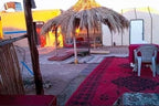 Desert Erg Chigaga Camp outdoor seating area with red and patterned rugs, low tables, and a thatched umbrella shade