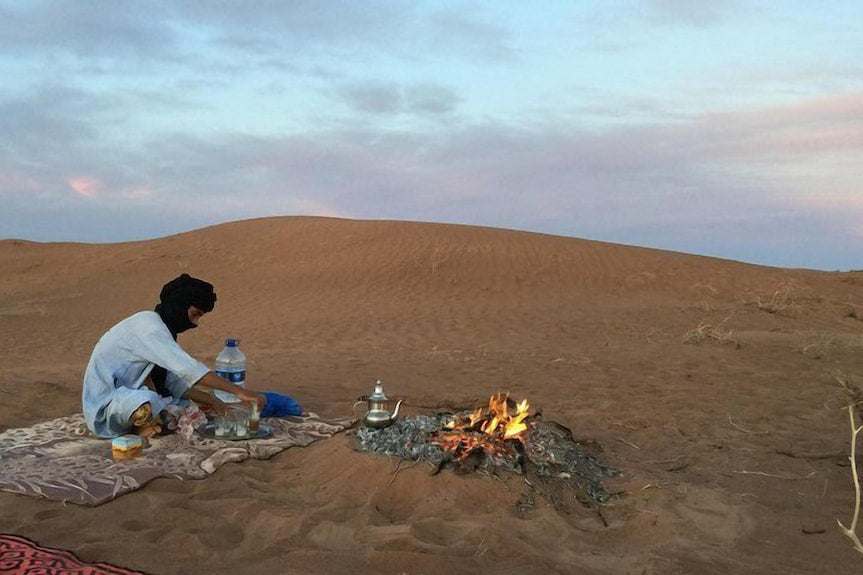 Desert Erg Chigaga Camp scene with person in traditional clothing preparing tea by a small campfire on sandy dunes under a soft sky