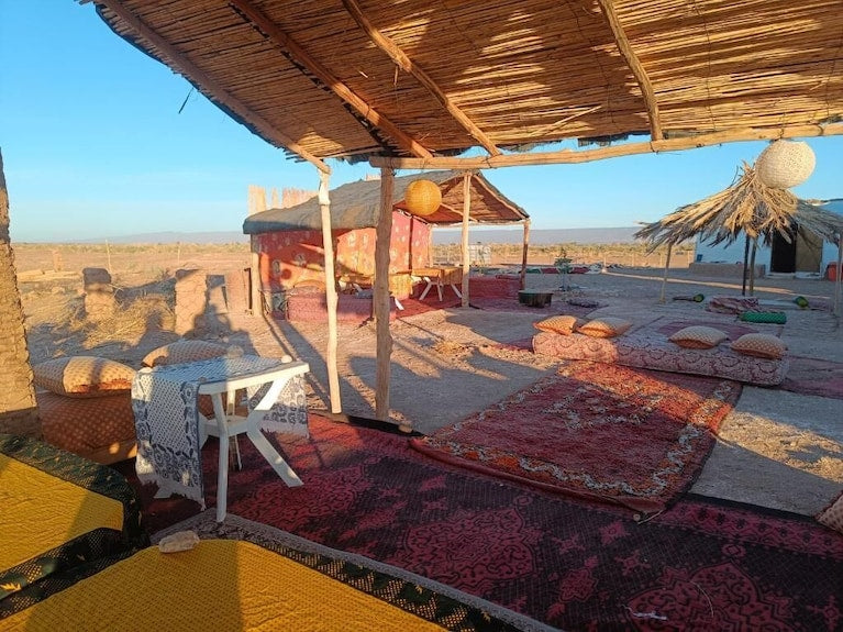 Desert Erg Chigaga Camp with a shaded seating area featuring wicker roof, colorful patterned rugs, cushions, and a small white table outdoors