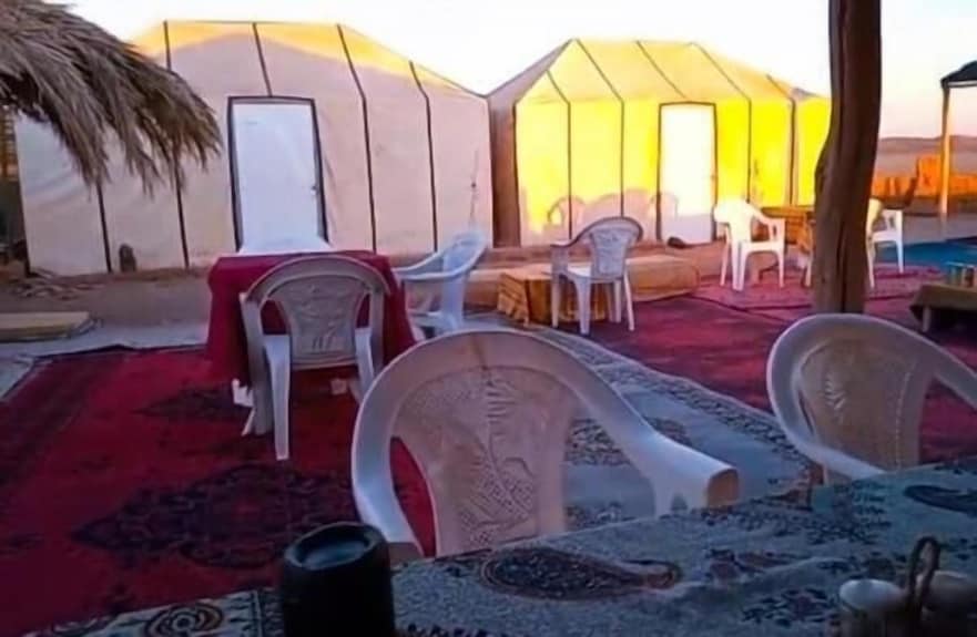 Desert Erg Chigaga Camp with traditional beige tents, plastic chairs, and tables on colorful patterned carpets under a shelter