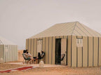 Beige fabric desert camp tents with vertical stripes and open doors, people sitting outside on chairs and a red patterned rug nearby during Desert Trip to Merzouga