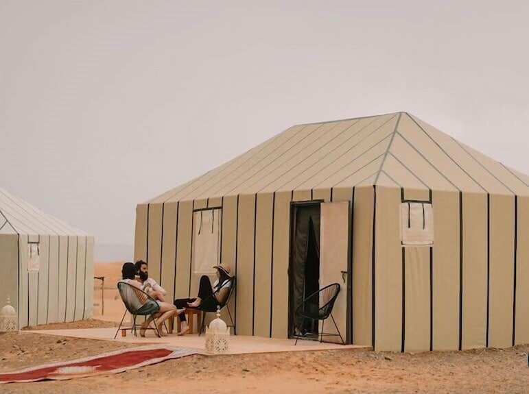 Beige fabric desert camp tents with vertical stripes and open doors, people sitting outside on chairs and a red patterned rug nearby during Desert Trip to Merzouga