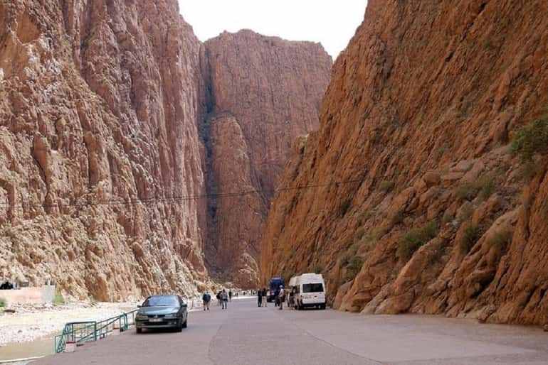 narrow paved road between tall reddish-brown rocky cliffs with parked cars and people walking during a Desert Trip to Merzouga