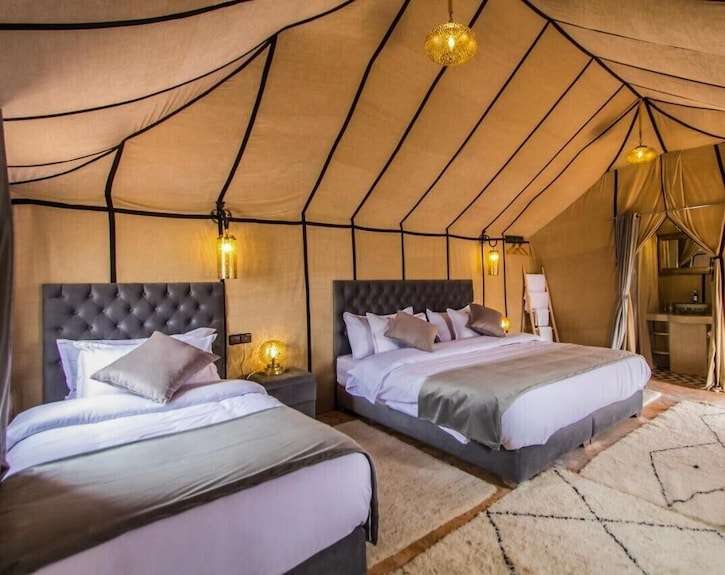 Desert Trip to Merzouga accommodation featuring a spacious beige tent interior with two double beds, gray tufted headboards, and soft ambient lighting