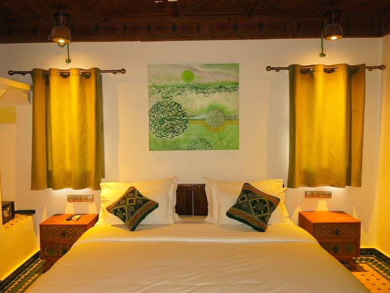 Desert Villa Boutique Hotel room with double bed, white linens, green patterned cushions, wooden nightstands, yellow curtains, and green artwork