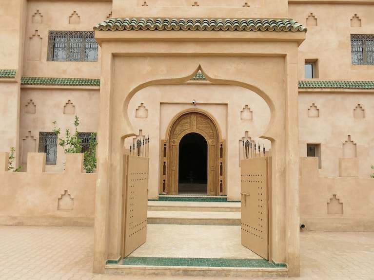 Desert Villa Boutique Hotel entrance with beige stucco walls, arched wooden door, and green tiled roof edge in Moroccan style