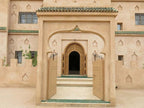 Desert Villa Boutique Hotel entrance with beige stucco walls, arched wooden door, and green tiled roof edge in Moroccan style