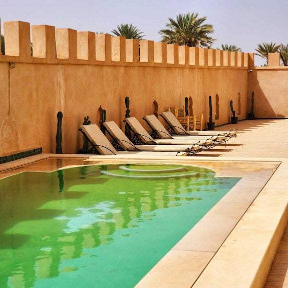 Desert Villa Boutique Hotel pool area with beige loungers lined against tan stucco walls under clear sky with palm trees
