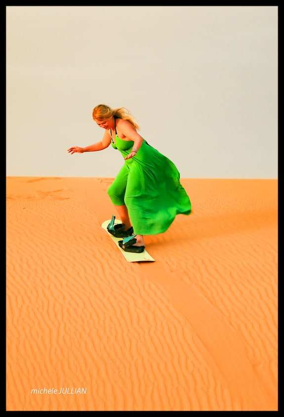 Desert Villa Boutique Hotel scene with a woman in a flowing green dress sandboarding on the orange desert dunes