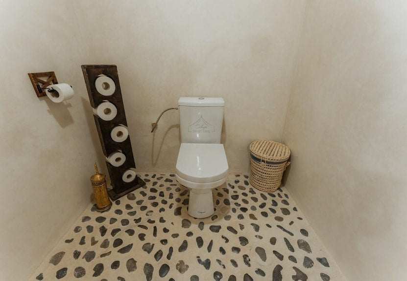 Dihya Desert Camp bathroom with white toilet, pebble-patterned floor tiles, woven basket, and wooden toilet paper stand