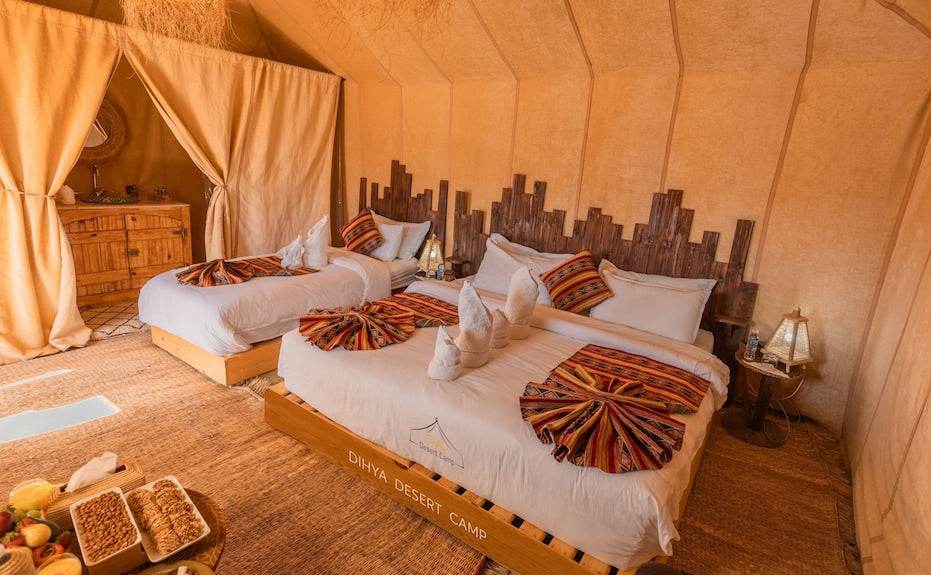 Dihya Desert Camp bedroom with two beds featuring white linens, patterned red and orange throws, wooden headboards, and beige fabric walls
