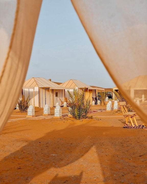 Dihya Desert Camp featuring beige canvas tents on orange desert sand with white lanterns and green plants under clear sky