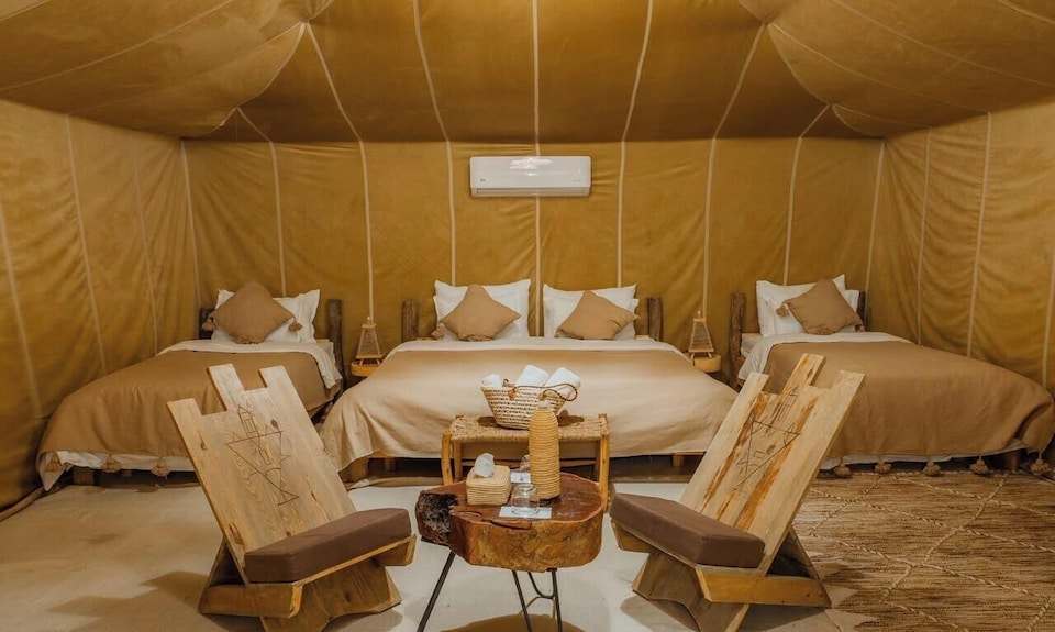 Dihya Desert Camp interior with beige fabric walls, three beds with tan bedding, and rustic wooden chairs with cushions