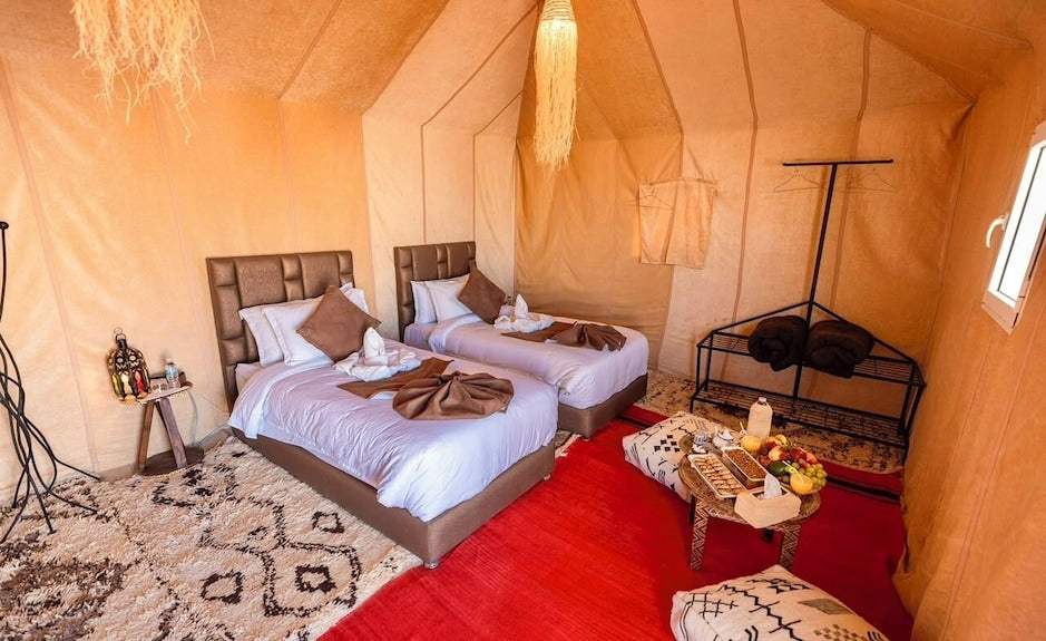 Dihya Desert Camp room with two brown leather headboard beds, white linens, patterned rugs, and a small table with fruit and snacks