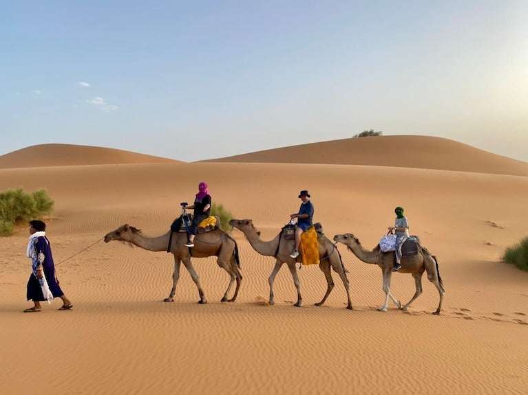 Dihya Desert Camp camel caravan with three riders in colorful traditional clothing crossing golden sand dunes under clear sky