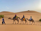 Dihya Desert Camp camel caravan with three riders in colorful traditional clothing crossing golden sand dunes under clear sky
