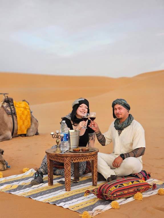Dihya Desert Camp desert picnic with couple wearing casual desert attire sitting on striped rugs, camel nearby