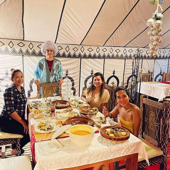 Dihya Desert Camp dining tent with four people enjoying traditional Moroccan dishes on a decorated table with patterned fabric walls