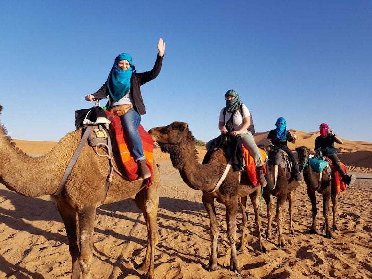 Dihya Desert Camp guests riding camels with colorful saddle blankets across sandy desert under clear blue sky