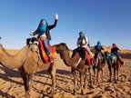 Dihya Desert Camp guests riding camels with colorful saddle blankets across sandy desert under clear blue sky