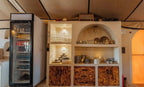 Dihya Desert Camp indoor kitchen area with glass-front refrigerator, wooden cabinets, shelves holding dishes, cups, and teapots