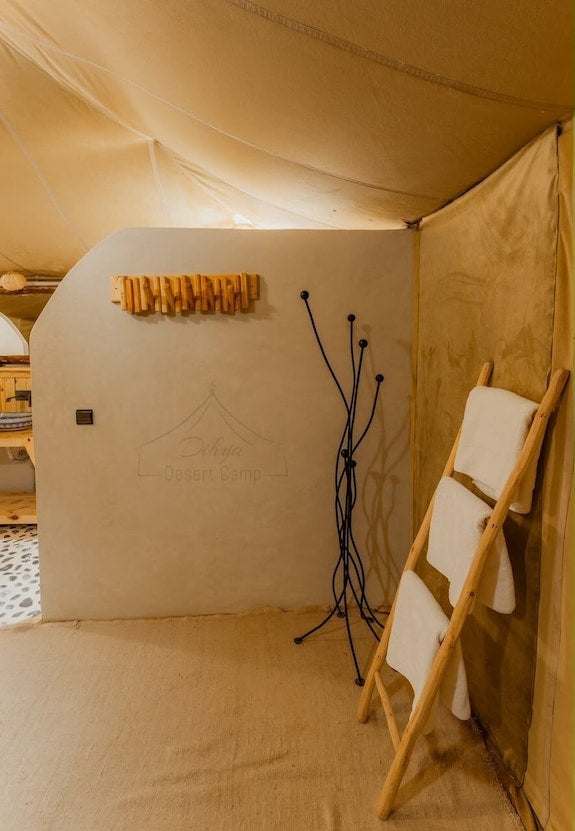 Dihya Desert Camp interior with beige tent fabric, smooth plastered wall, wooden towel rack, and black metal coat stand