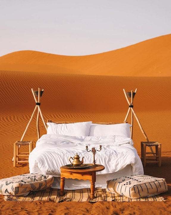 Dihya Desert Camp outdoor setup with white bedding on a double bed, wooden stools, patterned cushions, and a small wooden table in the desert sand
