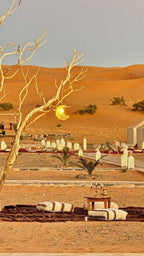 Dihya Desert Camp outdoor seating area with low wooden tables, cushions, lanterns, and desert sand dunes in background