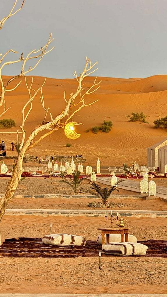 Dihya Desert Camp outdoor seating area with low wooden tables, cushions, lanterns, and desert sand dunes in background