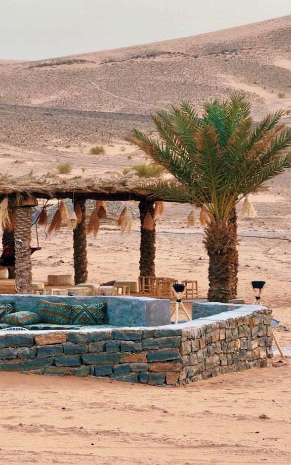 Dihya Desert Camp outdoor seating area with stone benches, patterned cushions, palm trees, and a shaded pergola in a desert landscape
