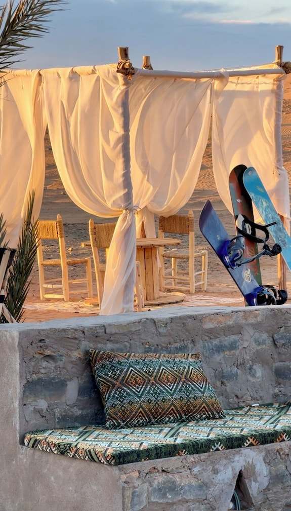 Dihya Desert Camp outdoor seating with flowing white curtains, wooden chairs, and patterned cushions on stone benches in desert