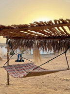 Striped fabric hammock under a rustic wooden pergola with woven fringe at Dihya Desert Camp in a sandy desert setting
