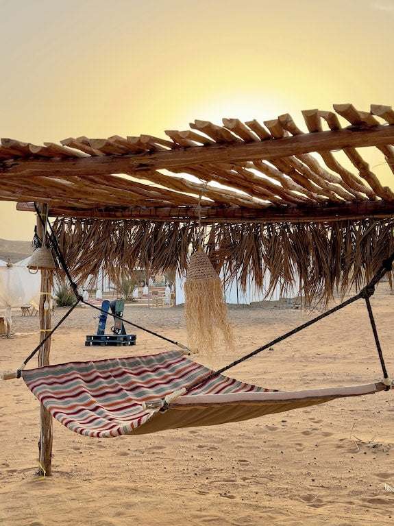 Striped fabric hammock under a rustic wooden pergola with woven fringe at Dihya Desert Camp in a sandy desert setting