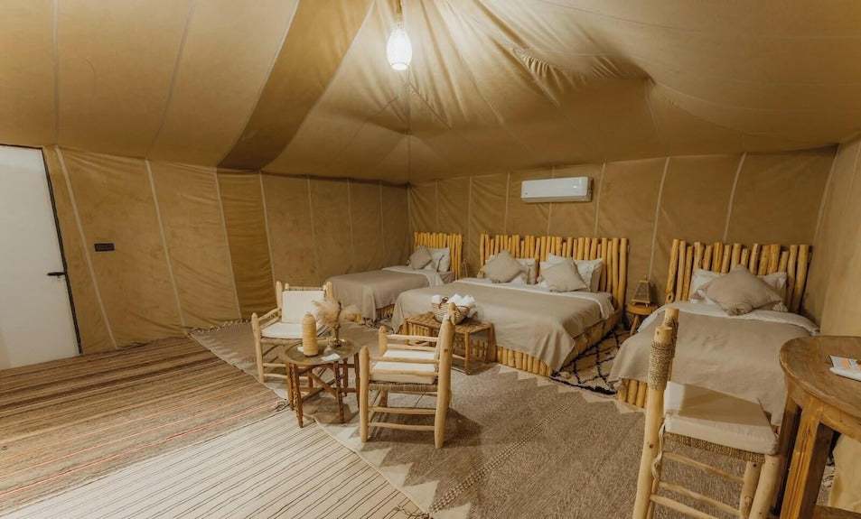 Dihya Desert Camp interior with three beds featuring bamboo headboards, beige linens, wooden chairs, and textured rugs