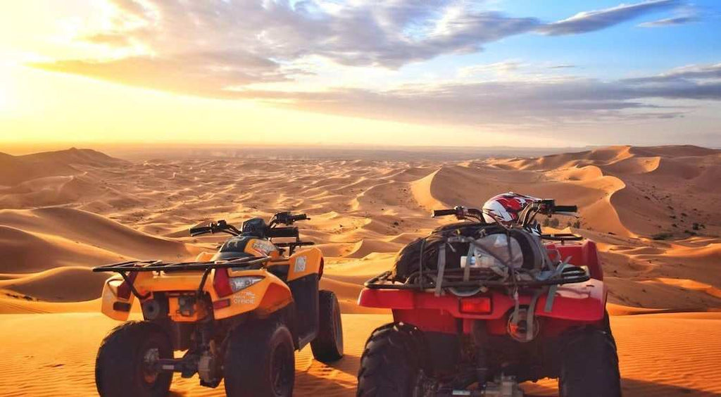 Two all-terrain vehicles, one orange and one red, parked on sandy dunes under a cloudy sky at Dihya Desert Camp