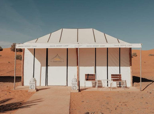 Dihya Desert Camp white canvas tent with tan trim set on sandy desert ground, featuring two wooden chairs and decorative lanterns