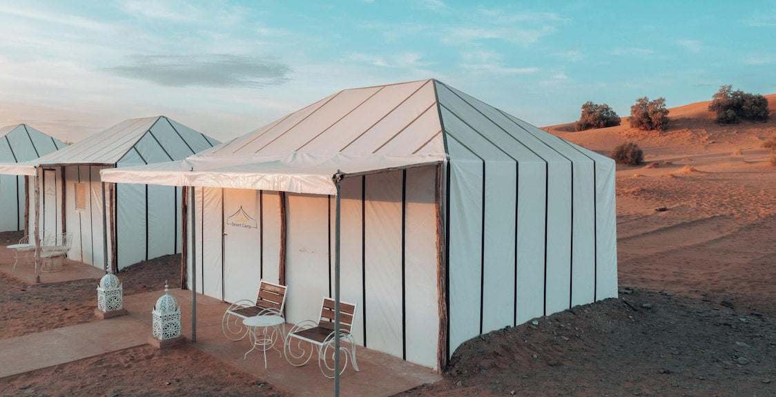 Dihya Desert Camp white canvas tents with pitched roofs and metal framed outdoor chairs in desert landscape during sunset