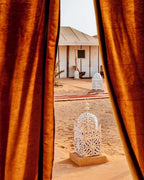 Dihya Desert Camp outdoor scene with white ornate lanterns on sandy ground framed by rust-colored curtains