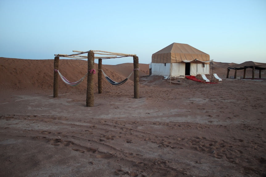 Erg Chigaga Star Camp with beige canvas tent, hammocks hanging on wooden posts, and sandy desert surroundings at dusk