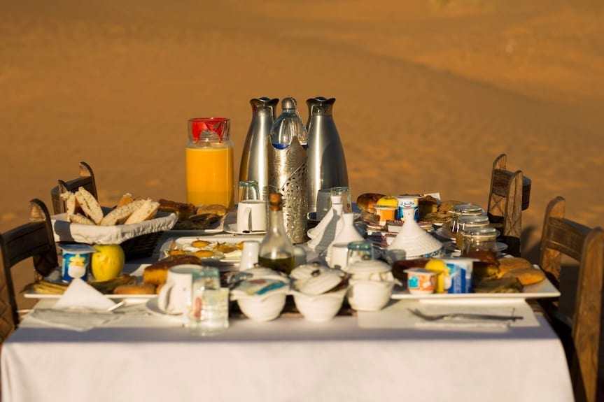 Erg Chigaga Star Camp outdoor breakfast spread with white tablecloth, bread, juice, cups, and metal thermoses in desert