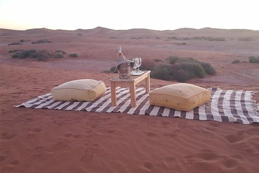 Erg Chigaga Star Camp striped outdoor rug with wooden table, glassware, wine bottle, and two yellow cushions on desert sand